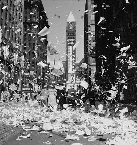 Black and white photograph. A Toronto Street, with a tall clock tower at the end. People crowd the street, barely discernible in the blizzard of tickertape paper in the air.